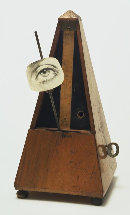 A wooden, brown, maybe oak or walnut-colored metronome. One the pendulum bar, there is a photographic paper cut-out, almost round but also a little square in shape, that displays a human eye. The image is a work of art in the style of Marcel Duchamp. It is a replica of the artwork titled 